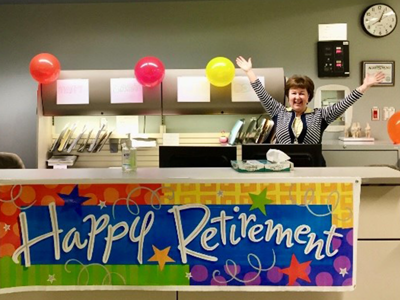 Donna Strauss behind a "Happy Retirement" banner, celebrating with outstretched arms high above her head.